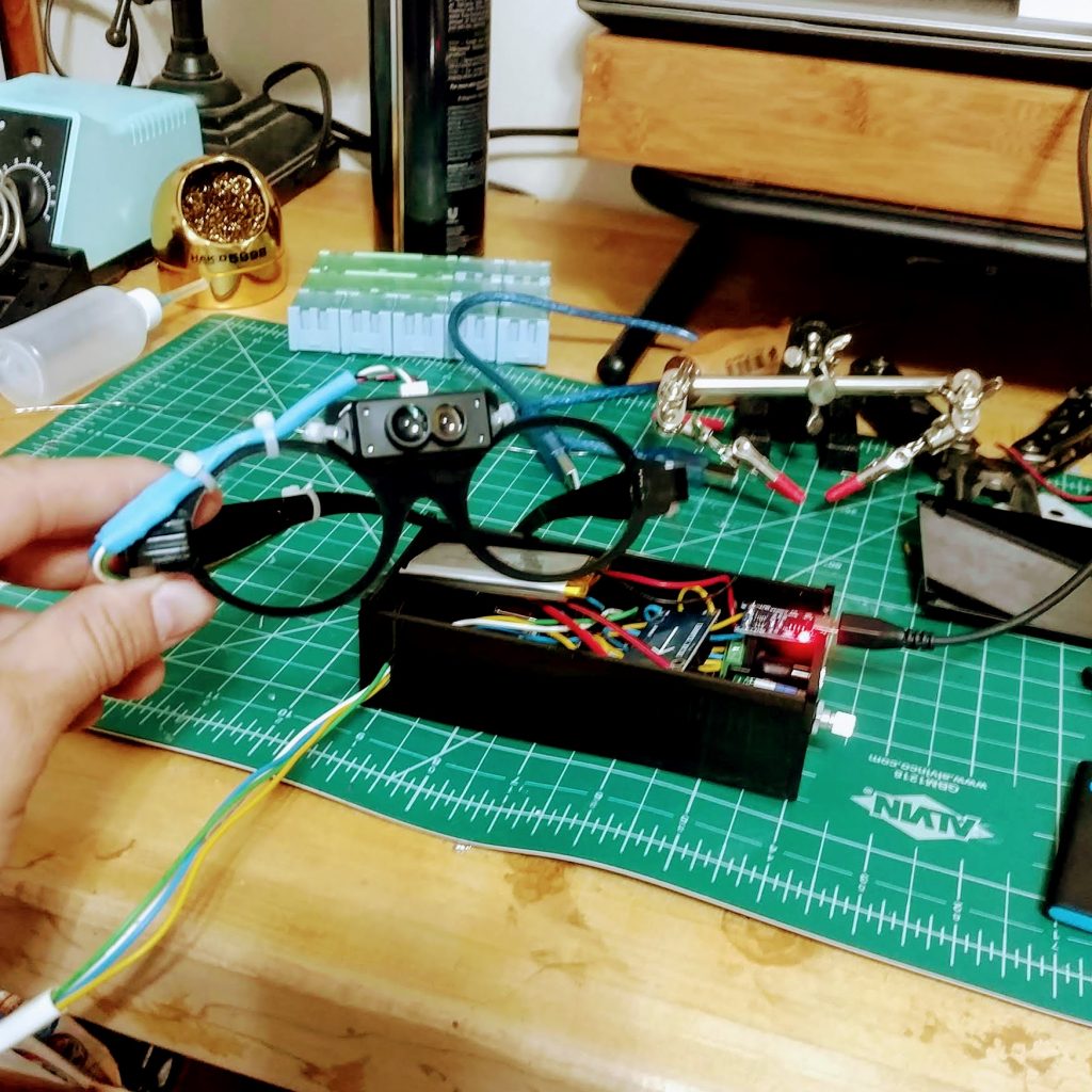 A LiDAR unit mounted to a pair of lasercut glasses with the control box in the background. 